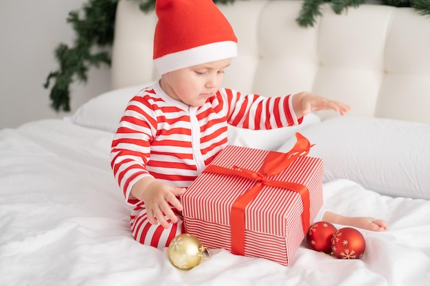 Dziewczynka w pasiastym body i czapce Mikołaja bawiąca się pudełkiem w świątecznej sypialni