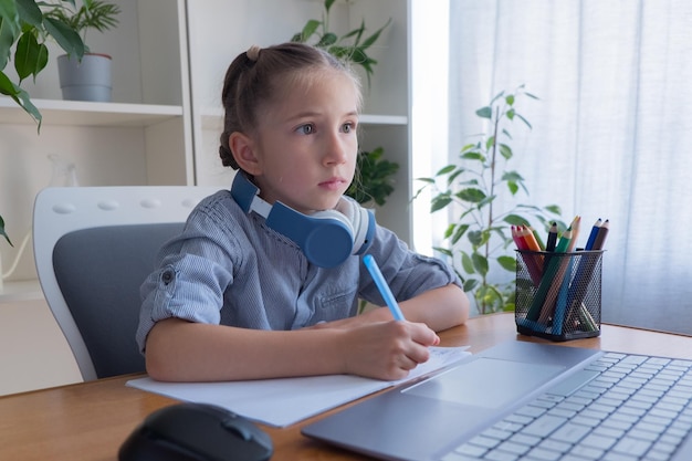 Dziewczynka w wieku 6-8 lat uczy się przy komputerze, odrabia prace domowe, komunikuje się przez internet, pisze w zeszycie.