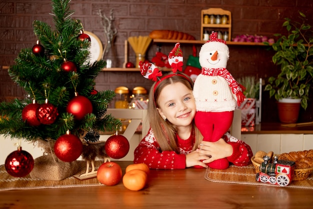 Dziewczynka z bałwanem w ciemnej kuchni w pobliżu choinki z czerwonymi kulkami raduje się i uśmiecha, koncepcja nowego roku i Bożego Narodzenia