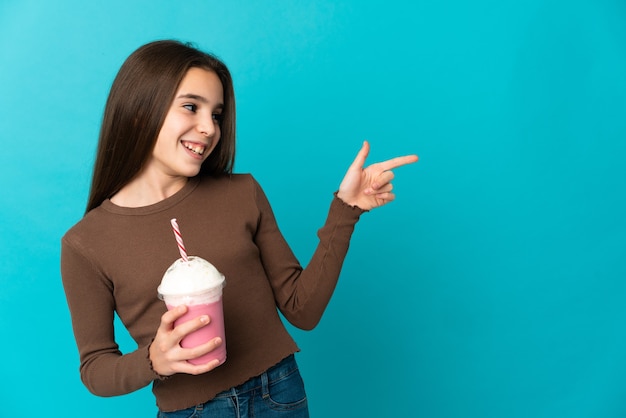 Dziewczynka z truskawkowym koktajlem mlecznym na białym tle na niebieskiej ścianie wskazując palcem na bok i prezentuje produkt