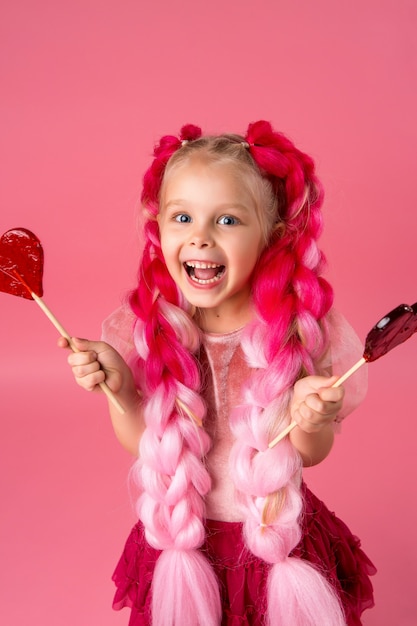 dziewczynka z warkoczami różowego kanekalonu trzyma Lollipopa w kształcie serca na różowym tle