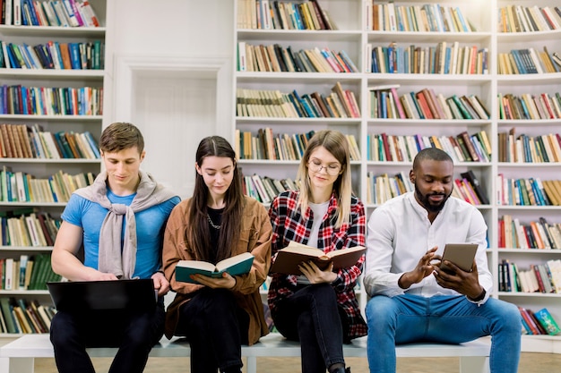 Dziewczyny I Chłopaki, Zapraszają Studentów Studiujących Razem W Bibliotece, Trzymających Tradycyjne Książki I Cyfrowe Tablety. Nauka Z Papierowymi Książkami I Gadżetami E-learningowymi.