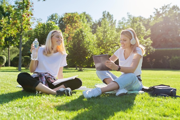 Dziewczyny nastoletnich studentów siedząc na zielonym trawniku w parku z plecakiem, książkami, cyfrowym tabletem, pić butelkę wody. Uniwersytet, kolegium, szkoła, edukacja i wiedza, styl życia nastolatków