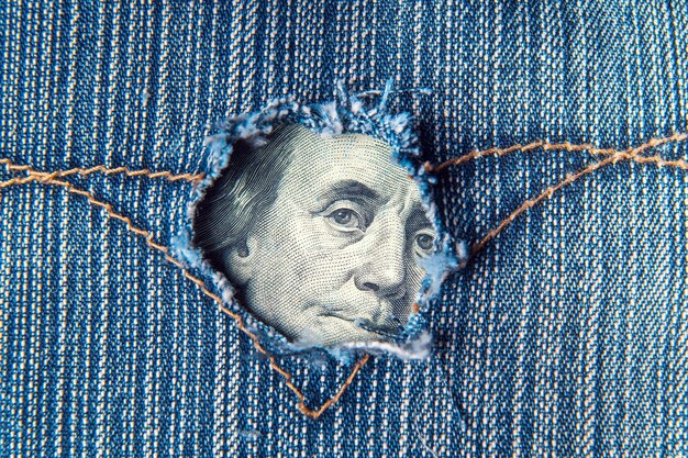 Dziura w kieszeni spodni dżinsowych, z której widać dolary Symbolizuje kryzys