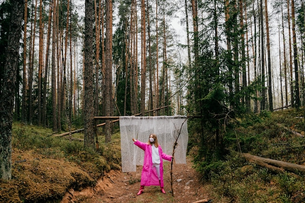 Dziwna młoda kobieta w masce i różowym płaszczu przeciwdeszczowym pozuje z kijem w lesie