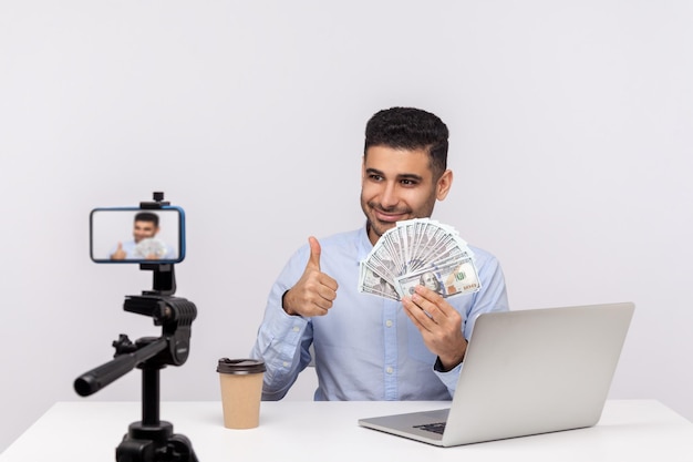 Edukacja finansowa Bogaty szczęśliwy biznesmen siedzi w miejscu pracy pokazując banknoty dolara i kciuk w górę nagrywania kursu, jak zarabiać pieniądze zysku kryty strzał studio na białym tle