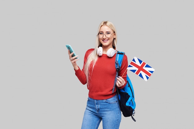 Edukacja online, tłumacz języka obcego, angielski, student - uśmiechnięta blond kobieta w słuchawkach trzyma telefon komórkowy i flagę brytyjską. Szara przestrzeń, kształcenie na odległość