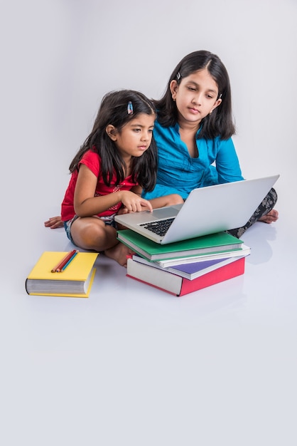 Edukacja w domu koncepcji - 2 Cute little zabawny indyjskich lub azjatyckich dziewcząt studiujących lub odrabiania lekcji na laptopie nad stosem książek, siedząc na podłodze w domu. Pojedynczo na białym tle