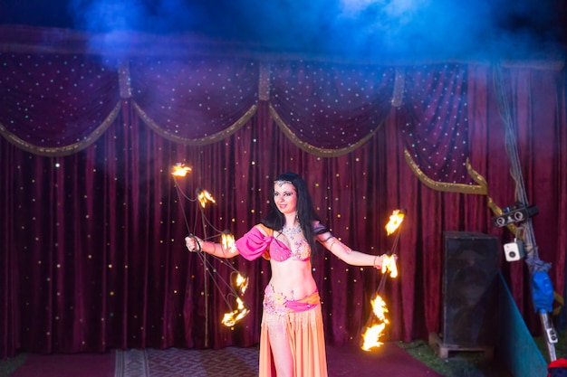 Egzotyczna ciemnowłosa tancerka ognia w jasnoróżowym i pomarańczowym kostiumie kręcąca się dwoma płonącymi pałeczkami na scenie z czerwoną kurtyną