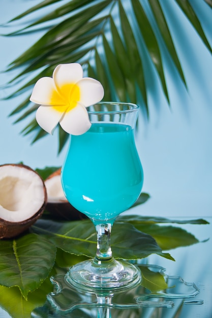 Egzotyczny tropikalny napój Blue Curacao koktajl w szklance z kwiatem Plumeria frangipani, liściem palmowym, świeżym kokosem na tle.