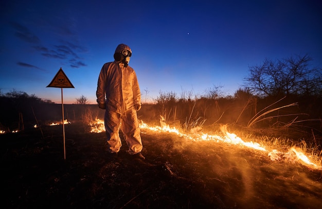 Ekolog gasi pożar w polu w nocy Mężczyzna w kombinezonie ochronnym i masce gazowej w pobliżu płonącej trawy z dymem i znakiem ostrzegawczym z czaszką i piszczelami Koncepcja klęski żywiołowej