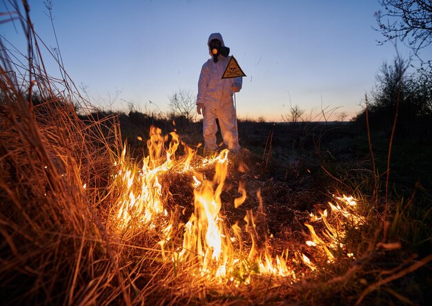 Ekolog strażak gasi pożar w polu w nocy Mężczyzna w garniturze i masce gazowej w pobliżu płonącej trawy z dymem trzyma znak ostrzegawczy z czaszką i piszczelami Koncepcja klęski żywiołowej