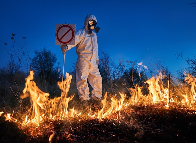 Ekolog strażak gaszący ogień na polu w nocy Człowiek w ochronnym garniturze przeciw promieniowaniu i masce gazowej w pobliżu płonącej trawy z dymem trzymający znak zakazu Koncepcja katastrofy naturalnej