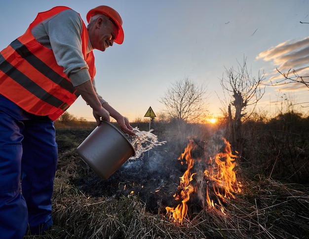 Ekolog strażak walczący z pożarem na polu z wieczornym niebem na tle Mężczyzna zajmujący się ochroną środowiska trzymający wiadro i wylewający wodę na płonącą suchą trawę w pobliżu znaku ostrzegawczego z znakiem wykrzyku