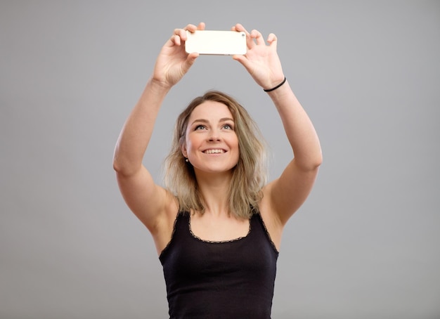 Ekspresyjna młoda kobieta robi zdjęcia przez telefon komórkowy na szarym tle