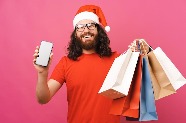 Ekstatyczny mężczyzna w świątecznym kapeluszu pokazuje ekran telefonu, trzymając torby z zakupami