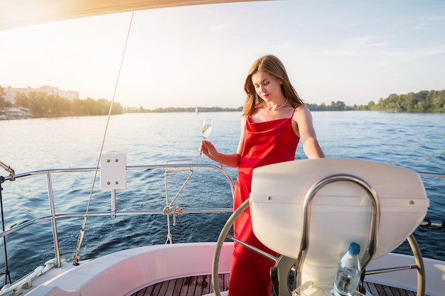 Elegancka atrakcyjna kobieta w długiej czerwonej sukience trzymająca kieliszek szampana stojąca na jachcie i patrząca na zachód słońca nad rzeką