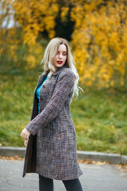 Elegancka blondynka w szarym płaszczu pozuje w jesiennym parku
