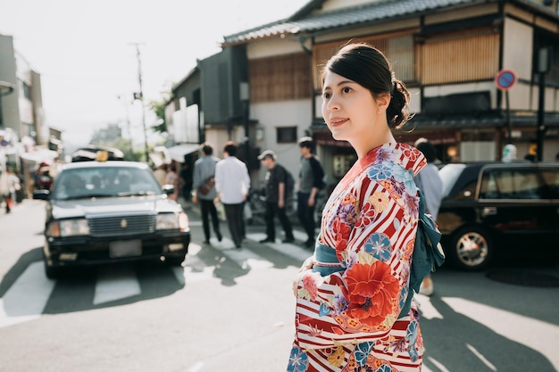 elegancka dama azjatyckich w kolorowe tkaniny kimono chodzenie na drodze z samochodami ruchliwymi jazdą. piękna Japonka w tradycyjne tkaniny szczęśliwy patrzeć na zachód słońca na ulicy. miejscowi mężczyźni na przejściu dla pieszych.