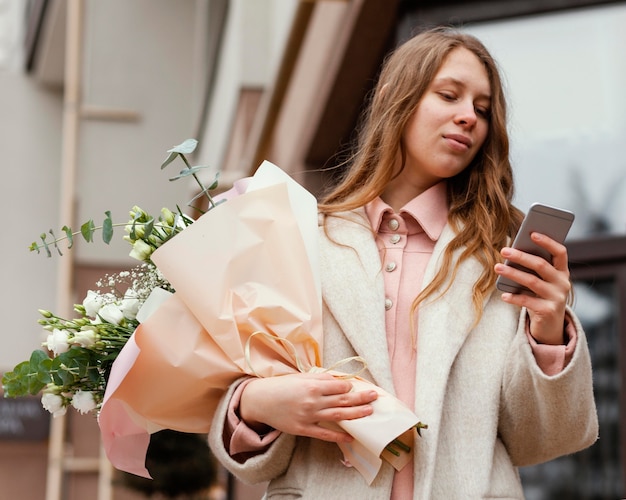 Zdjęcie elegancka kobieta trzyma bukiet kwiatów na zewnątrz i za pomocą smartfona
