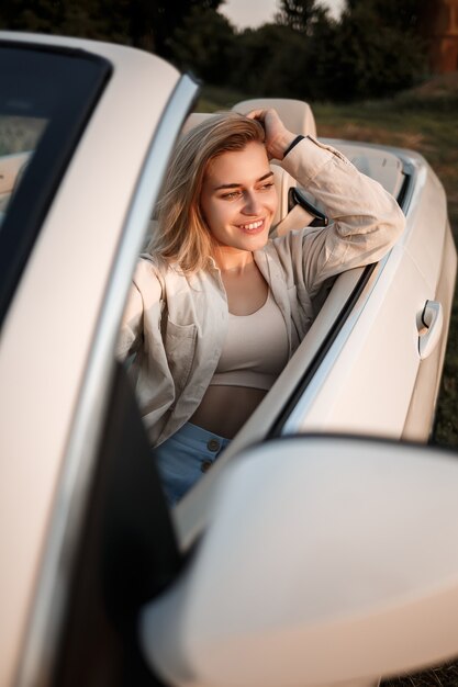 Elegancka luksusowa dziewczyna o blond włosach, uśmiechnięta siedząc na białym kabriolecie. Młoda, odnosząca sukcesy kobieta siedzi w swoim białym samochodzie