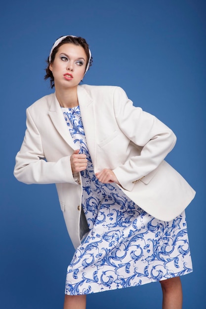 elegancka młoda kobieta w białej marynarce, wzorzystej sukience, butach, szaliku na głowie pozuje na niebieskim tle