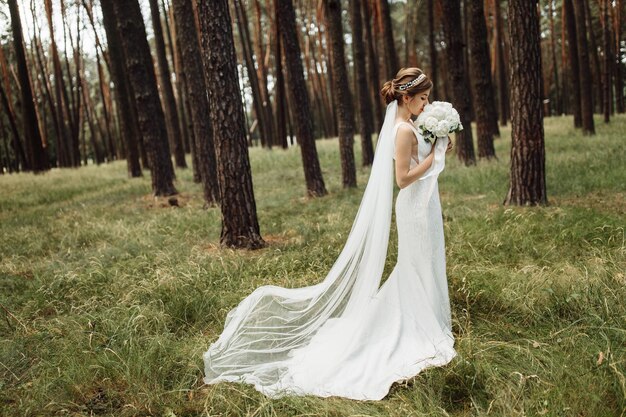 Elegancka panna młoda stoi w lesie i trzyma piękny bukiet ślubny z piwonią w dłoniach