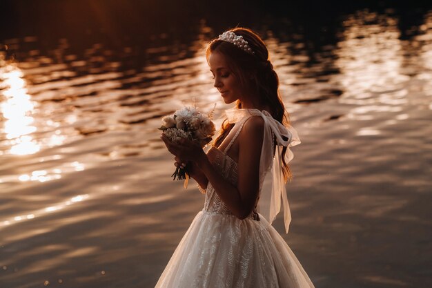 Elegancka panna młoda w białej sukni i rękawiczkach stoi nad rzeką w parku z bukietem, ciesząc się naturą o zachodzie słońca.Modelka w sukni ślubnej i rękawiczkach w parku przyrody.Białoruś