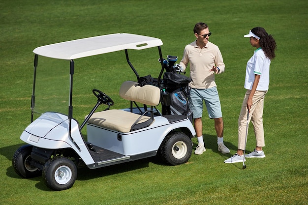 Elegancka para stojąca przy wózku golfowym na zielonym polu i rozmawiająca w słońcu