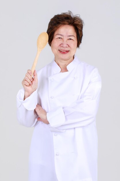 Elegancka starsza kobieta jako profesjonalny szef kuchni pewnie stojący z ramieniem skrzyżowanym i drewnianą łyżką i widelcem w kuchni restauracji. Starsza ciocia ekspertka w gotowaniu dla zdrowej kuchni osoby na emeryturze.