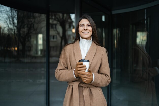 Elegancka uśmiechnięta kobieta z filiżanką kawy na tle szklanych drzwi biurowca