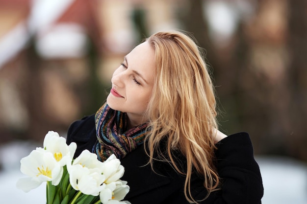 Zdjęcie elegancka uśmiechnięta młoda kobieta o długich blond włosach w czarnym płaszczu w mieście kobieta z kwiatami