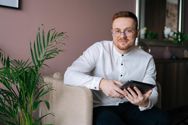 Elegancki, atrakcyjny uśmiechnięty mężczyzna w modnych okularach, siedzący w miękkim fotelu i korzystający z komputera typu tablet w domu, patrząc w kamerę