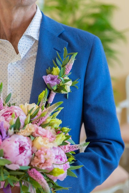 Elegancki mężczyzna w garniturze trzyma okrągły bukiet z piwoniami i eustomą Butonierka z białej eustomy eremurus i zieleni przymocowana do niebieskiej kurtki pana młodego Szczegóły ślubu i florystyka
