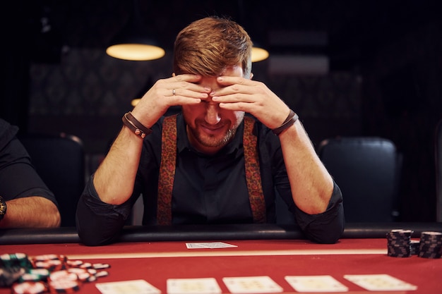 Elegancki młody mężczyzna siedzi w kasynie i źle się czuje, ponieważ przegrywa w pokera