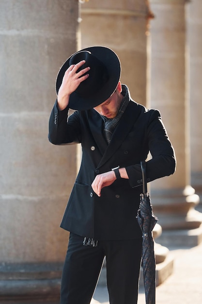 Elegancki młody mężczyzna w czarnym kapeluszu i ubraniach z parasolem w rękach na zewnątrz w mieście