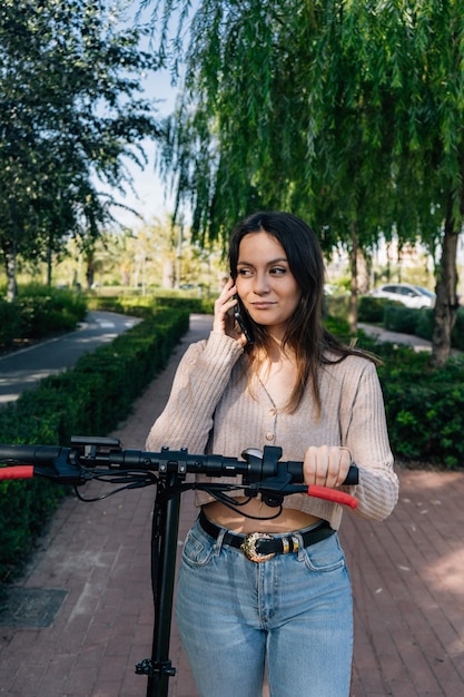 Elektryczna hulajnoga i urządzenie mobilne Dziewczyna rozmawia ze swoim smartfonem podczas spaceru z elektryczną hulajnogą obok niej przez park miejski