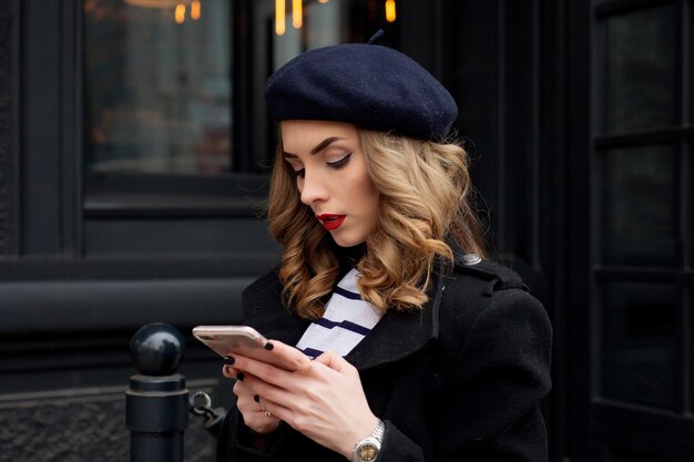 Emocje, ludzie, piękno i styl życia koncepcja - dziewczyna Francuzka. Street photo młoda kobieta nosi stylowe ubrania klasyczne. Koncepcja moda kobiece. Styl francuski. Dziewczyna wysyłająca SMS-y na smartfonie