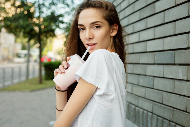 Emocje, styl życia, uroda, ludzie, koncepcja żywności-miejski portret młodej atrakcyjnej kobiety brunetka w białej koszulce picia koktajl mleczny przez rurkę lato o zachodzie słońca