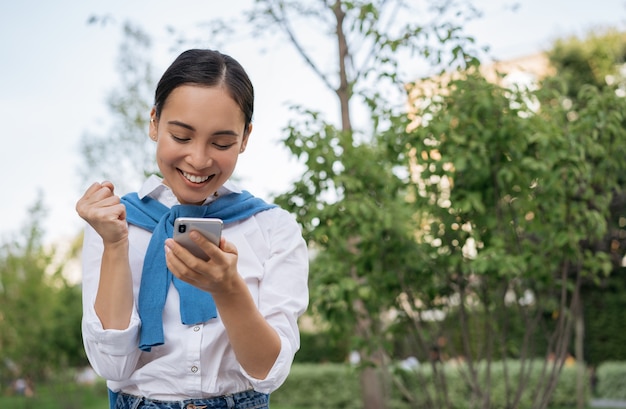 Emocjonalna Azjatka korzystająca z telefonu komórkowego, patrząc na cyfrowy ekran, wygrywająca loterię online, świętowanie sukcesu