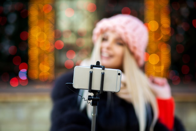 Emocjonalna blondynka w różowej czapce, biorąc selfie na ulicy ozdobionej girlandami. Miejsce na tekst