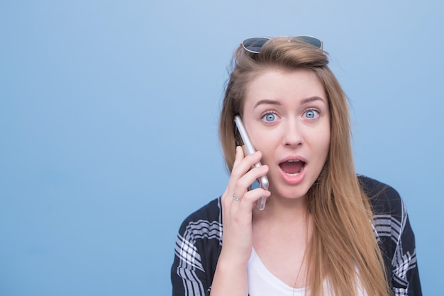 Emocjonalna dziewczyna opowiada na telefonie i patrzeje kamerę na błękitnym tle.