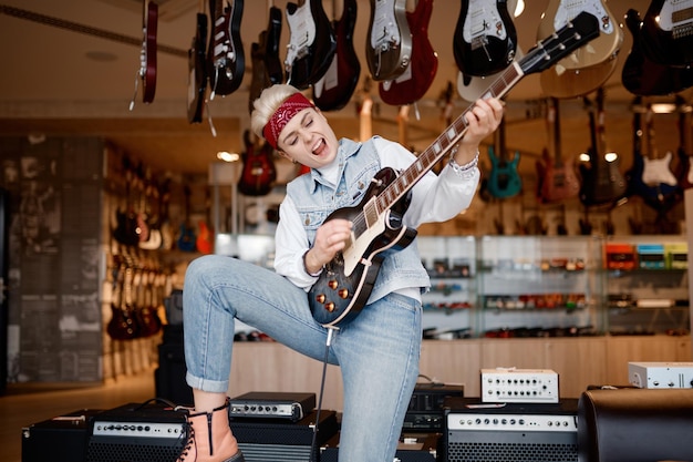Emocjonalna hipsterska rockmanka grająca na gitarze podłączona do wzmacniacza dźwięku stereo, czująca satysfakcję i podziw dla dźwięku