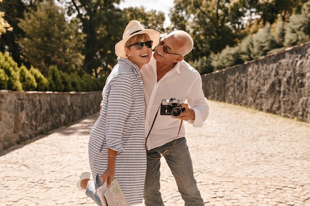 Emocjonalna Kobieta Z Krótką Blond Fryzurą W Długiej Pasiastej Bluzce I Kapeluszu śmiejącym Się Z Mężczyzną Z Wąsami W Koszuli Z Kamerą W Parku