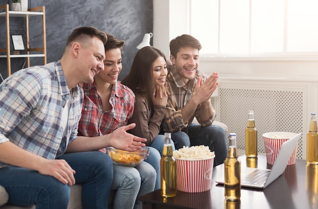 Emocjonalni przyjaciele oglądają film na laptopie, jedzą popcorn i piją piwo w domu, w pomieszczeniu