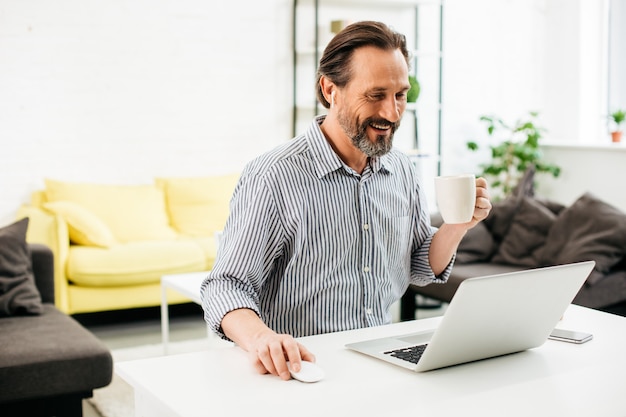 Emocjonalny brodaty mężczyzna siedzi przy stole z filiżanką kawy i uśmiecha się, patrząc na ekran nowoczesnego laptopa
