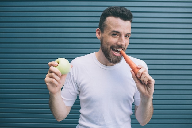 Emocjonalny mężczyzna w białej koszuli stoi. Uśmiecha się i pozuje. Facet gryzie marchewkę, a drugą trzyma zielone jabłko. Na białym tle na paski.