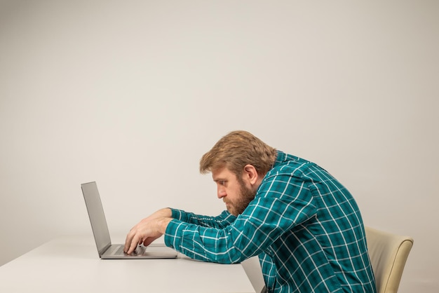 Emocjonalny mężczyzna z brodą w pozycji zgarbionej, siedzący w pokoju biurowym, pracujący z laptopem