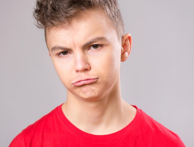 Emocjonalny portret białego, zdenerwowanego, problematycznego nastolatka, smutnego chłopca patrzącego w kamerę, zaniepokojonego dziecka w czerwonej koszulce na szarym tle.