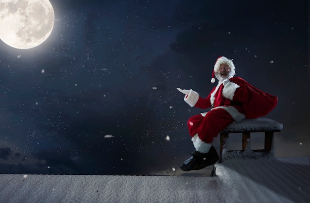 Emocjonalny Święty Mikołaj gratulujący Nowego Roku 2021 i Bożego Narodzenia. Człowiek w tradycyjnym stroju siedzi na dachu domu z pełni księżyca na tle w północy. Zima, wakacje, wyprzedaże. Miejsce.